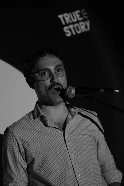 Un uomo con gli occhiali racconta al microfono, sotto la luce di scena.